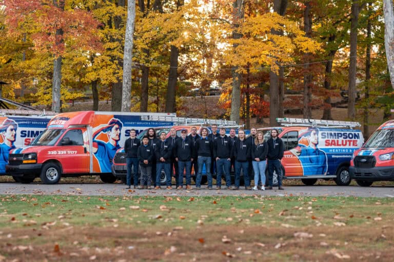 A group of people stands in front of three Simpson Salute service vans parked on a tree-lined street with autumn foliage.
