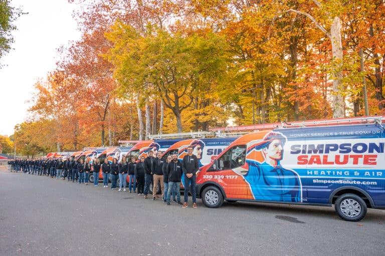 A line of people in uniform stand beside several Simpson Salute Heating & Air vans, parked in front of trees with autumn foliage.