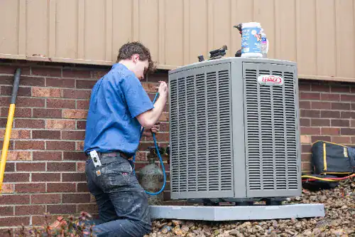 A technician in a blue shirt kneels beside an outdoor air conditioning unit, using tools to perform maintenance or repairs.