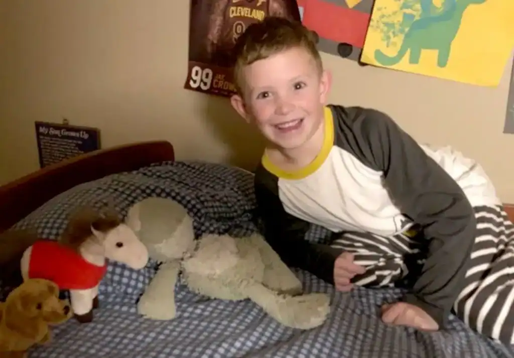 A young boy in pajamas smiles while sitting on a bed with a plaid blue blanket, surrounded by stuffed animals, under posters on a beige wall.