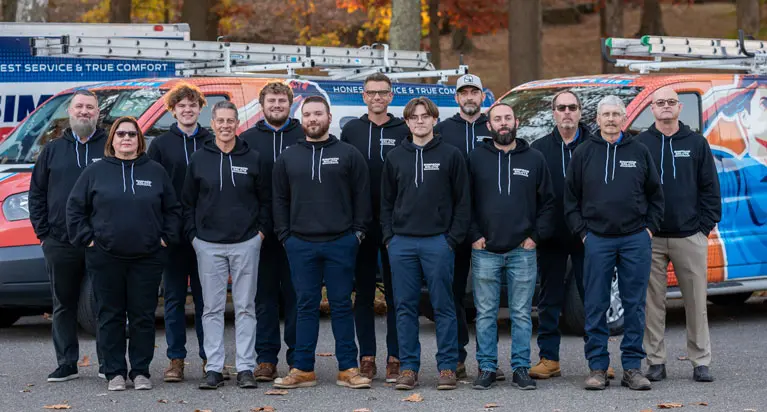 A group of thirteen people in matching black hoodies stands in front of two work vans outdoors on a paved surface with trees in the background.